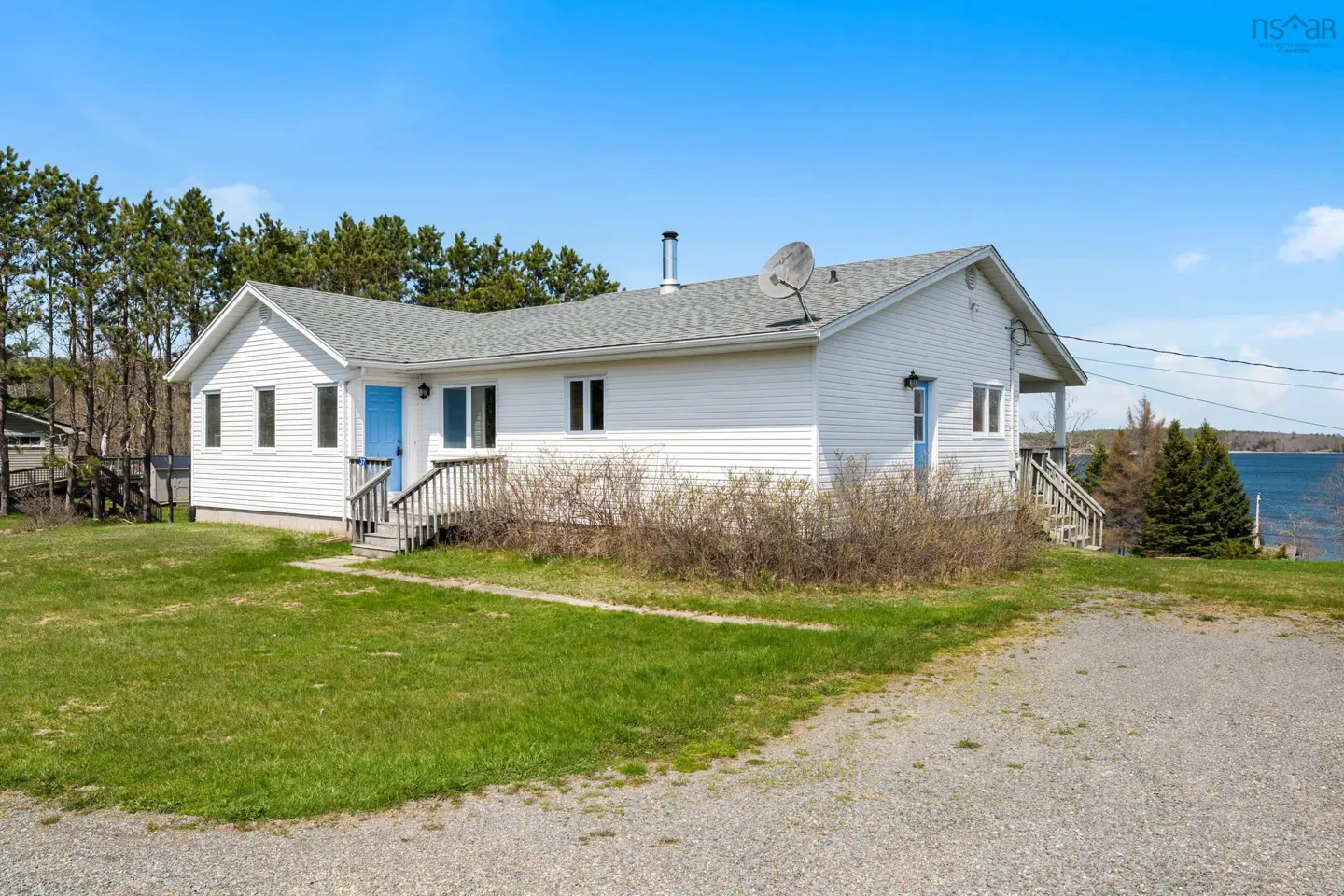 A white, one-story house with a blue door and gray roof sits on a green lawn under a blue sky.