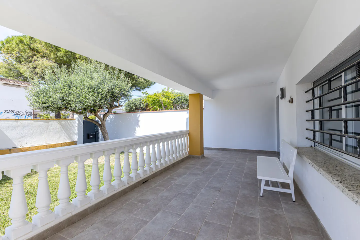 Covered patio with white balustrade, gray tile floor, and white bench. A tree and lawn are visible beyond the railing.