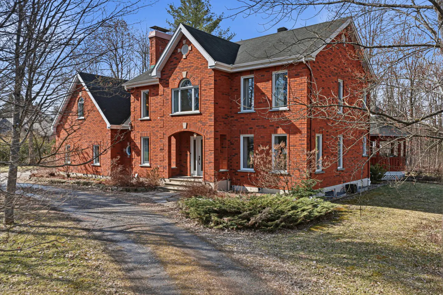 Two-story red brick house with a black roof, white trim, and a chimney. A driveway leads to the front entrance. Trees surround the property.