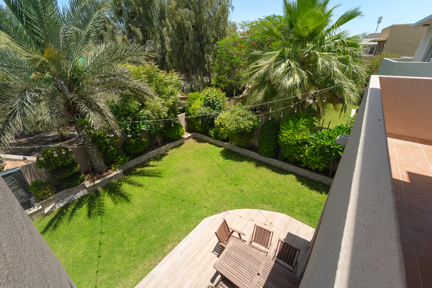 Overhead view of a green backyard with palm trees, a wooden deck, and a table with chairs. String lights hang above the yard.