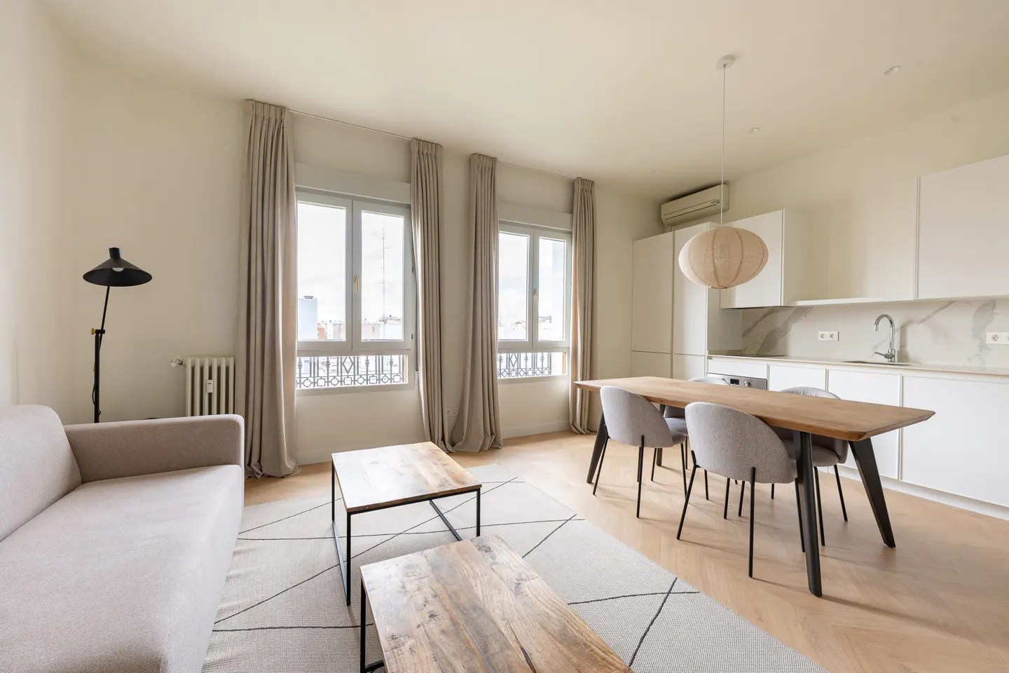 Bright, modern apartment interior with a beige sofa, wooden coffee tables, dining table with chairs, and a white kitchen. Two windows with beige curtains.