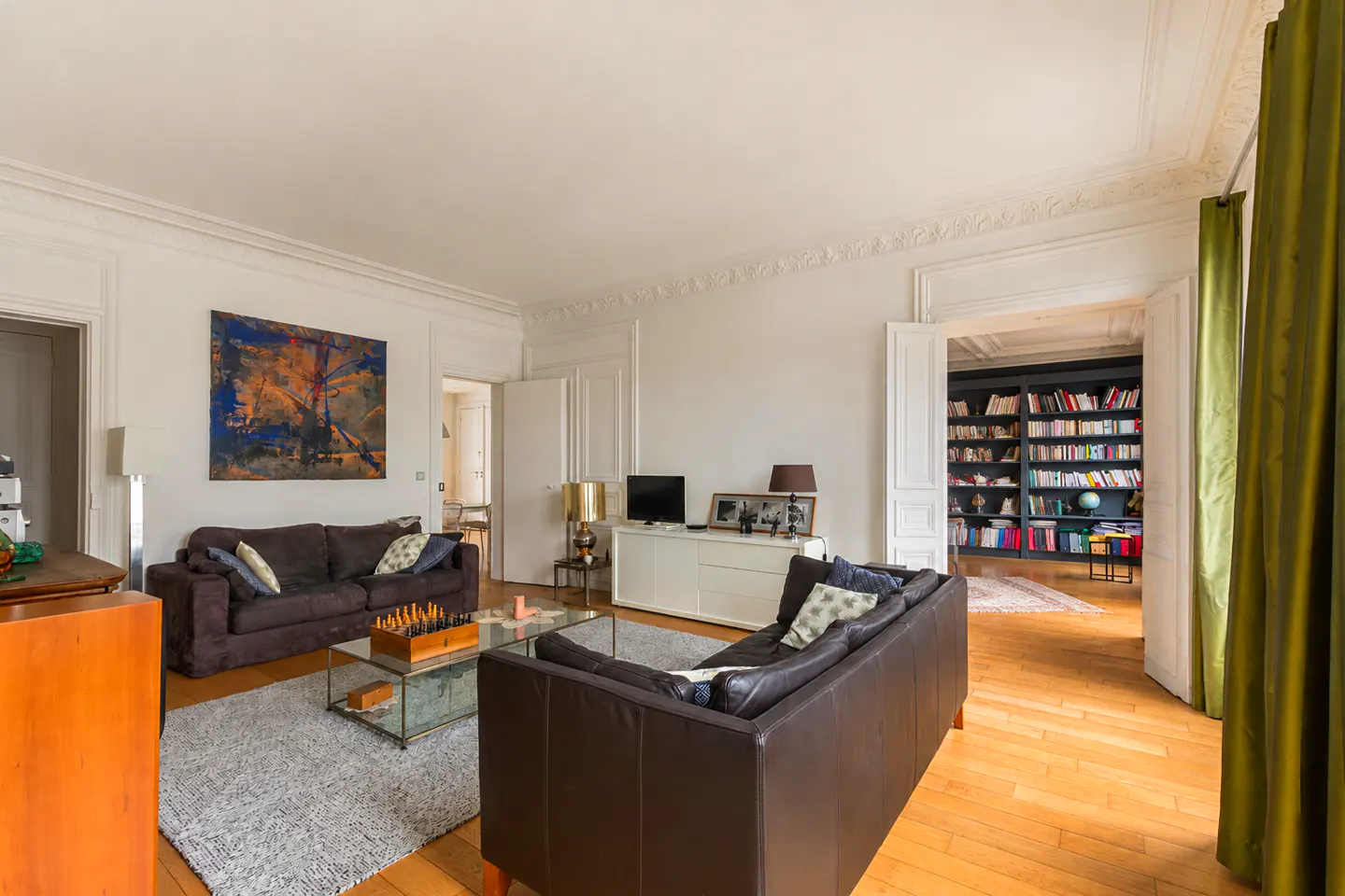 Bright living room with hardwood floors, two brown sofas, and a glass coffee table with a chess set. A bookcase is visible through an open doorway.