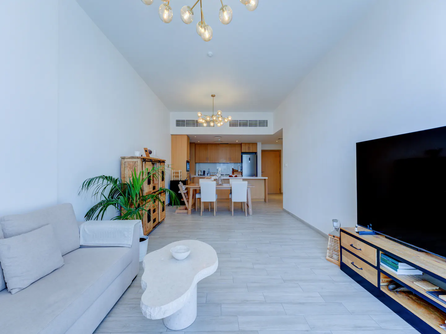 Bright, open-concept living space with a gray sofa, unique white coffee table, and a large TV. The kitchen and dining area are visible in the background.