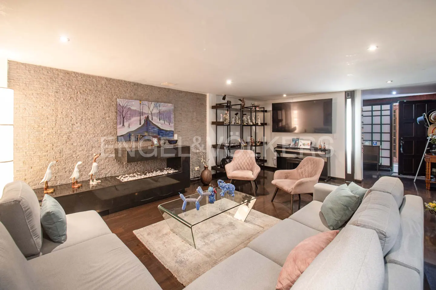 Living room with gray sofa, glass table, pink chairs, and a black fireplace with white stones. A painting hangs above the fireplace.
