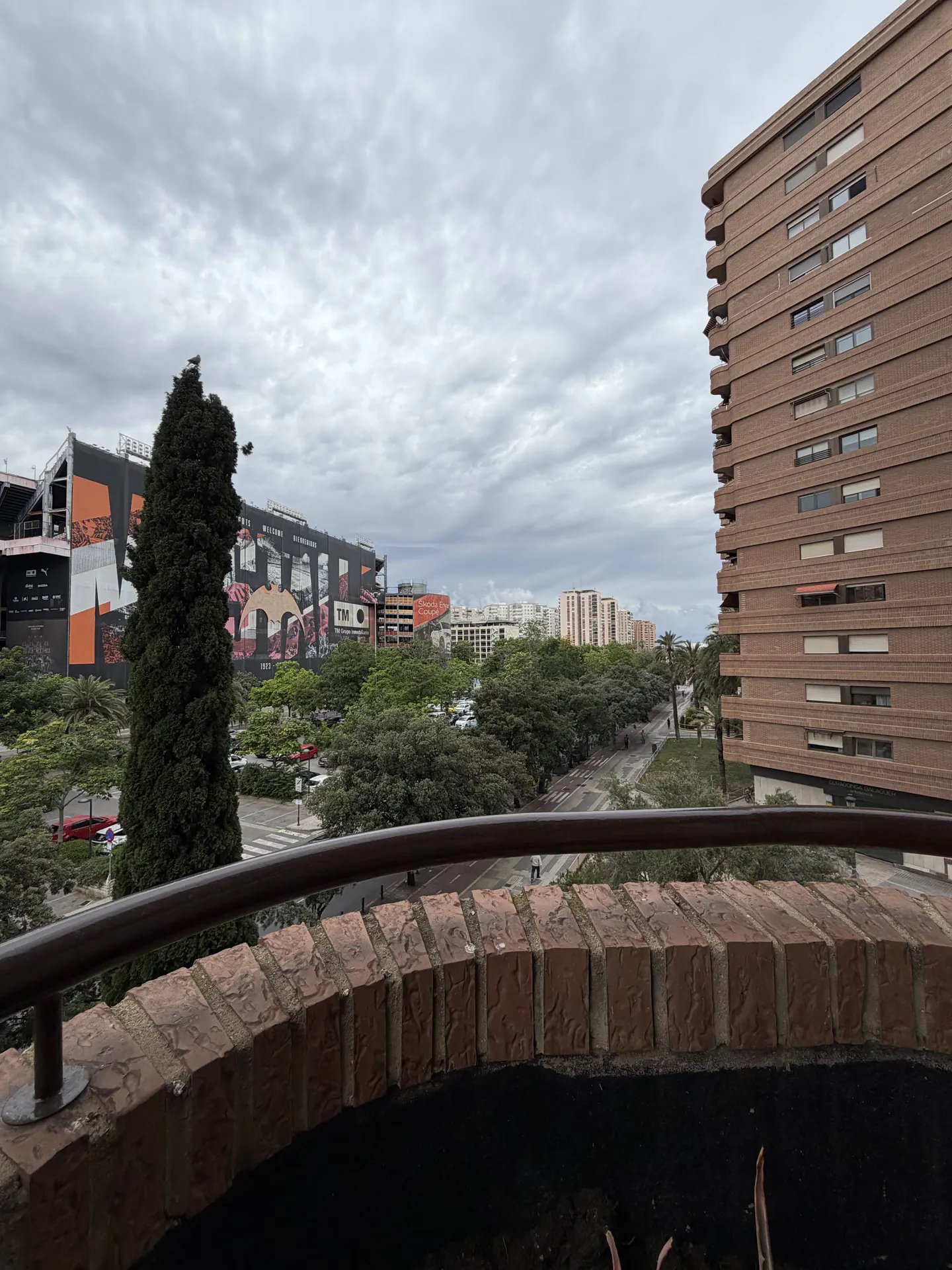 View from a balcony with a brick railing, overlooking a city street, trees, and a stadium with colorful murals under a cloudy sky.
