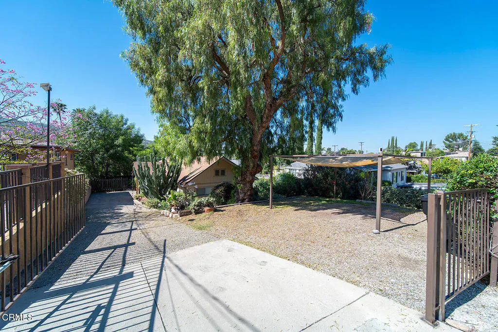 A sunny backyard with a large tree, gravel, and a covered patio area. A fence and gate are visible in the foreground.