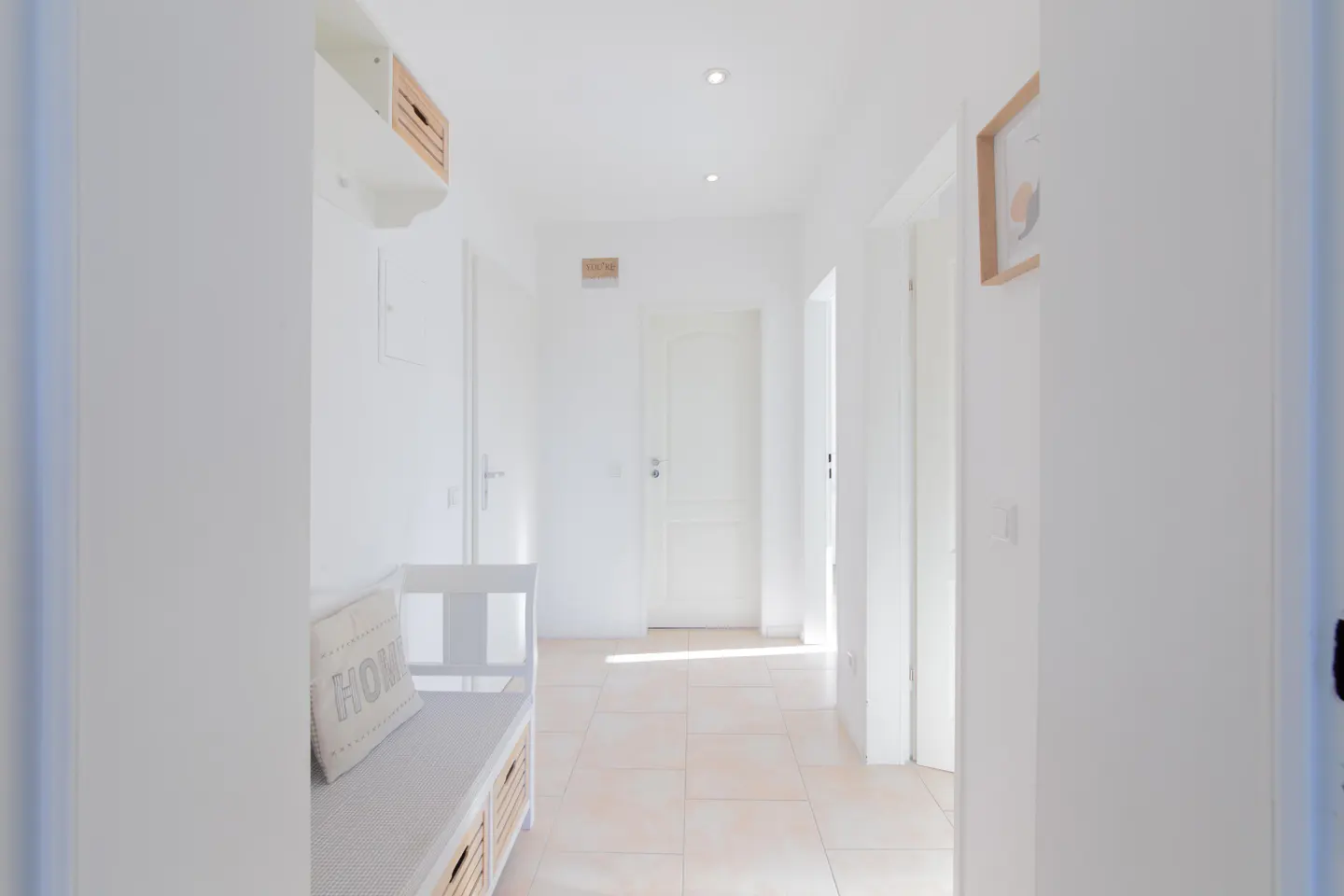 Bright hallway with white walls and tile floor. A white bench with a "HOME" pillow sits to the left. Doors line the hallway.