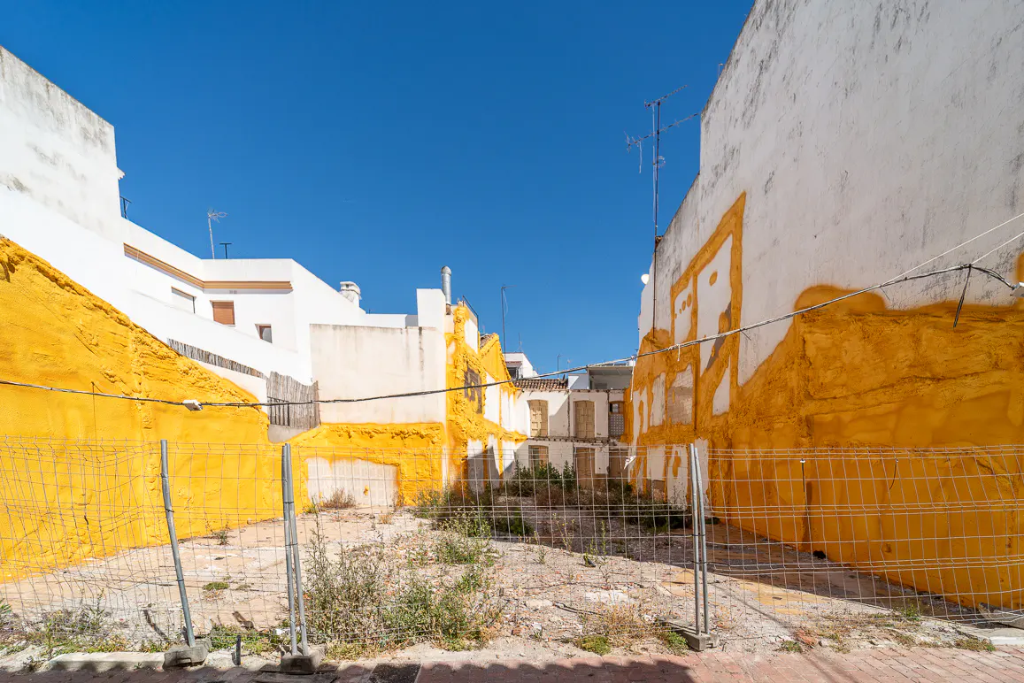 Vacant lot with white buildings and yellow paint, under a clear blue sky. A wire fence encloses the overgrown area.