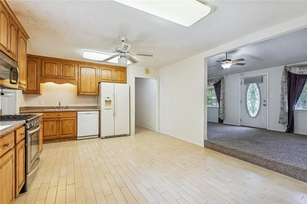 A bright kitchen with wood cabinets, white appliances, and light wood floors, leading to a carpeted living room.