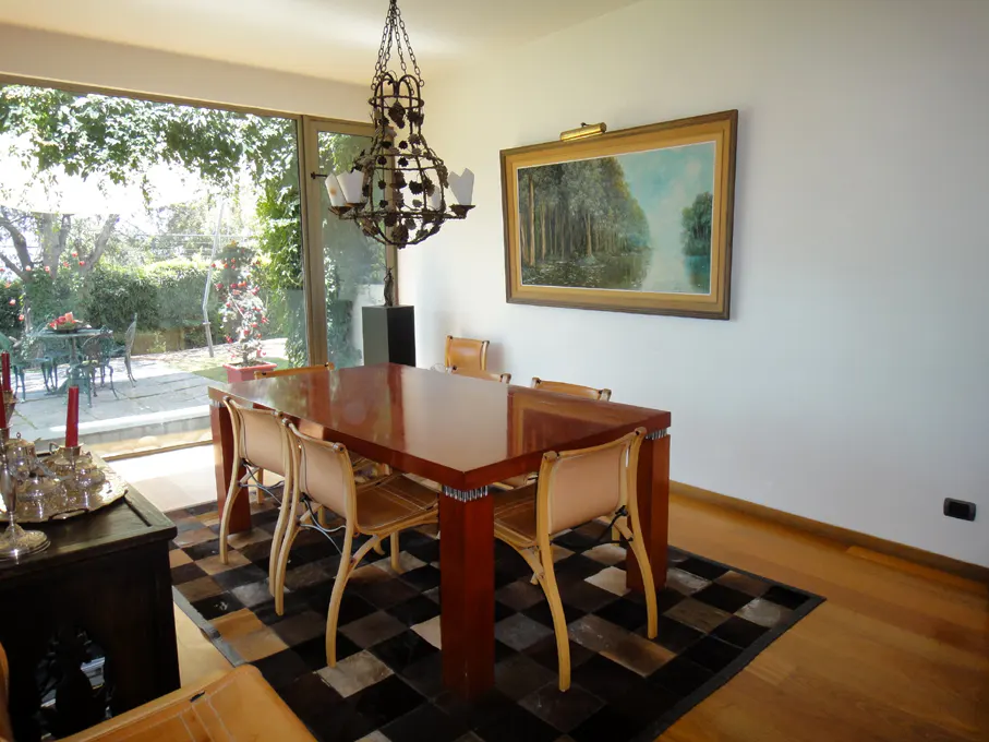 A dining room with a wood table, tan chairs, and a black and tan checkered rug. A chandelier hangs above the table. A painting is on the wall.