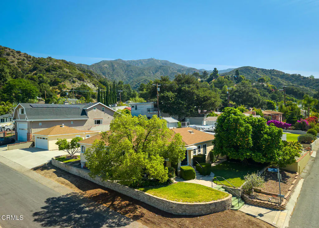 Aerial view of a yellow house with a brown roof, surrounded by green trees and a stone wall, with mountains in the background.