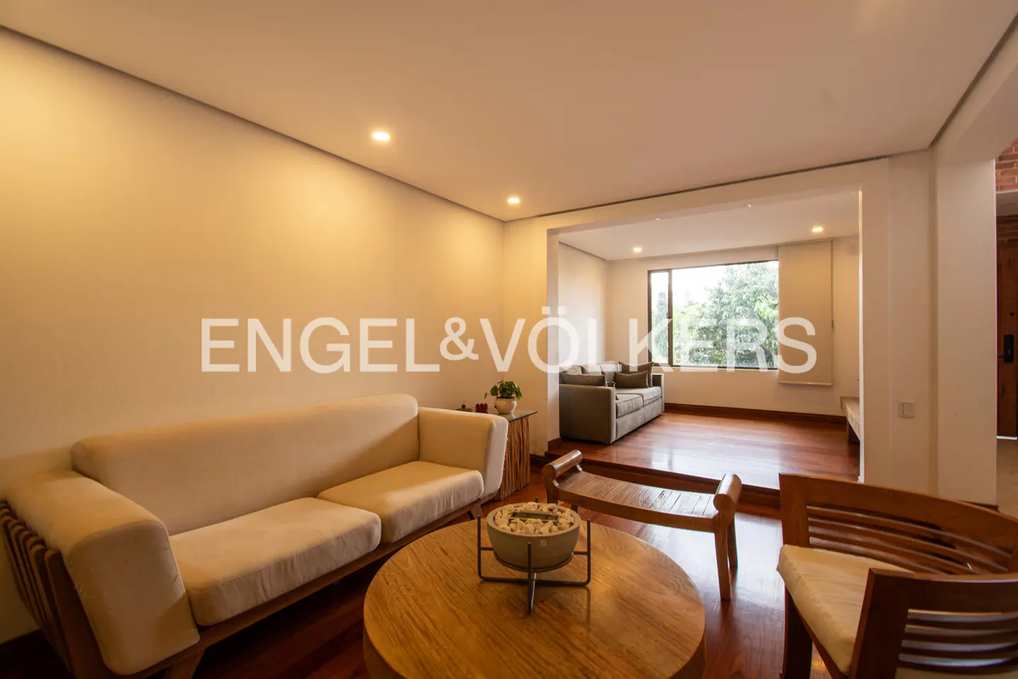 Living room with a beige sofa, wooden chairs, and a round wooden table on a hardwood floor. A second living area is visible through an opening.