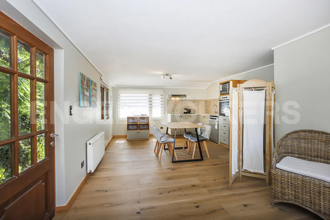 Open-concept living space with wood floors, a dining table, and a small kitchen. A wooden door leads to a green outdoor view.