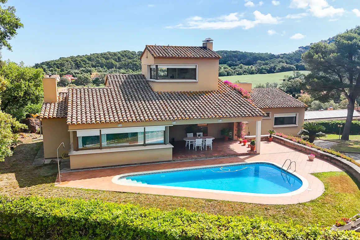 Tan house with a red tile roof, a blue pool, and green landscaping on a sunny day.