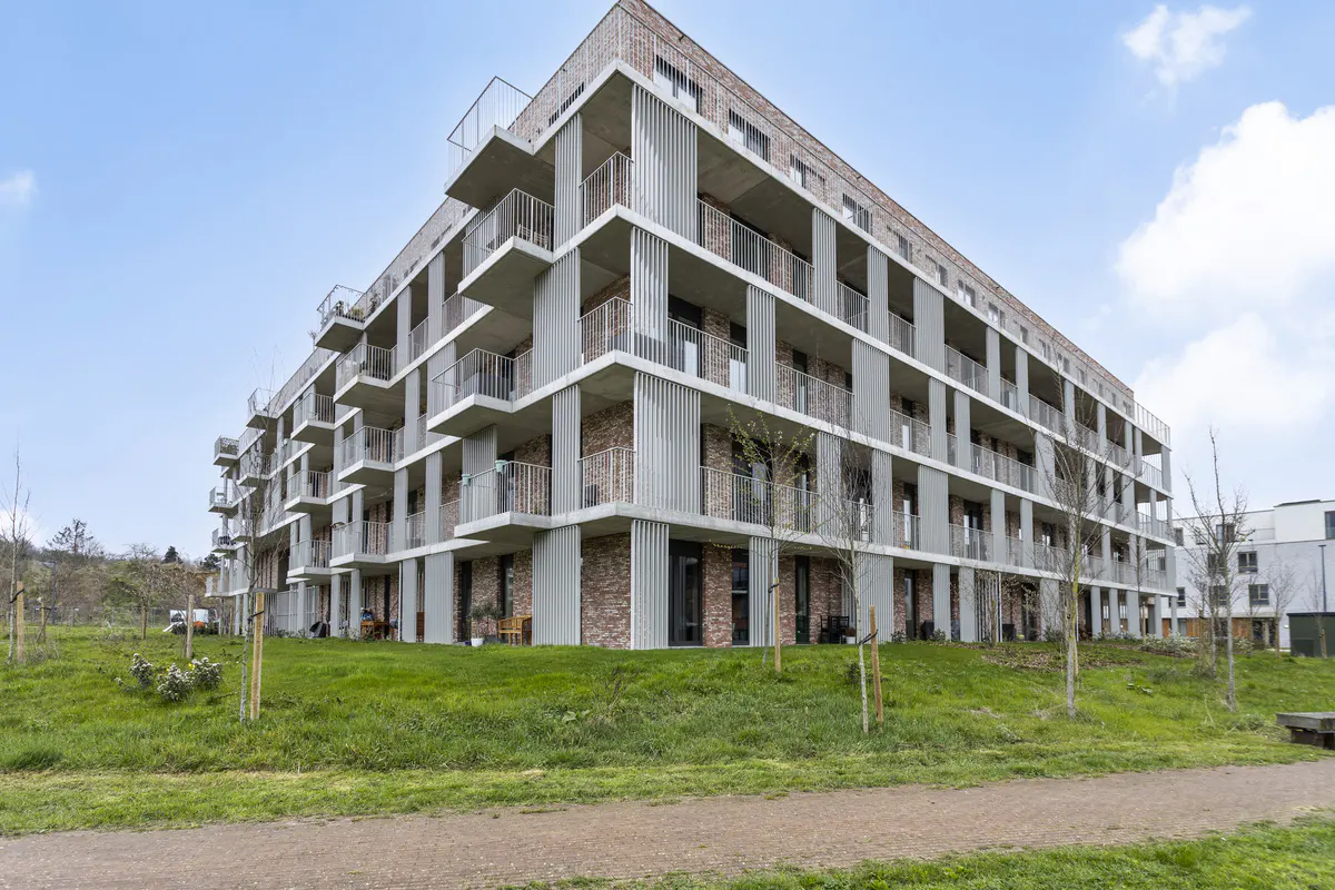 Modern apartment building with balconies, gray concrete and brick facade, surrounded by green lawn and trees under a blue sky.