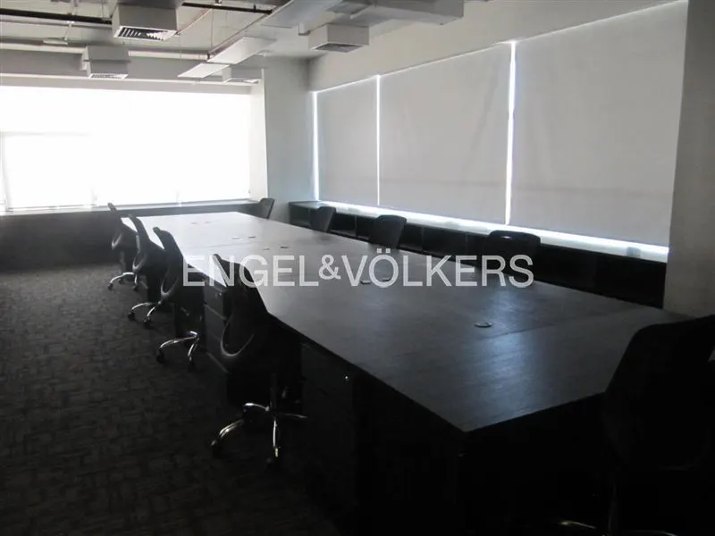 A long, dark conference table with black chairs in a bright, modern office space with white window shades.