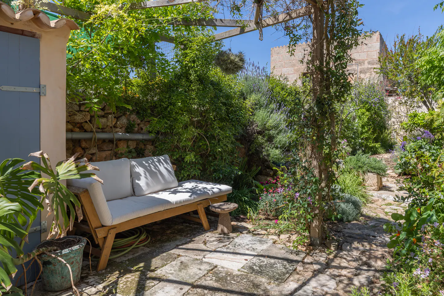 Outdoor patio with stone flooring, a wooden bench with white cushions, and lush green plants. A blue door is visible on the left.