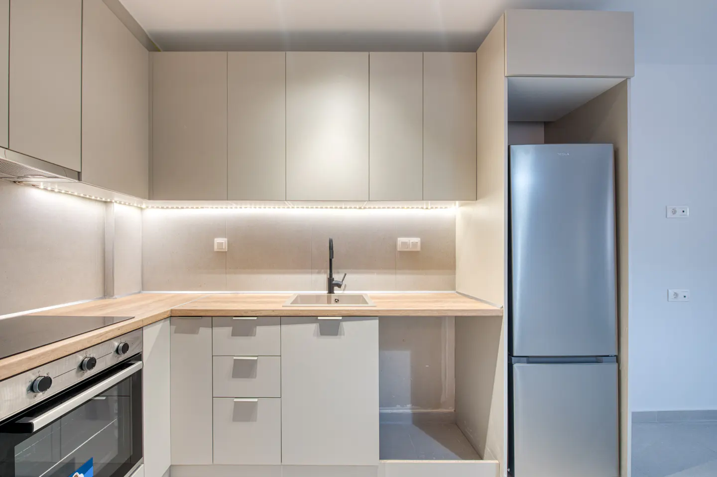 A modern kitchen with light gray cabinets, wood countertops, and stainless steel appliances. Under-cabinet lighting illuminates the sink area.