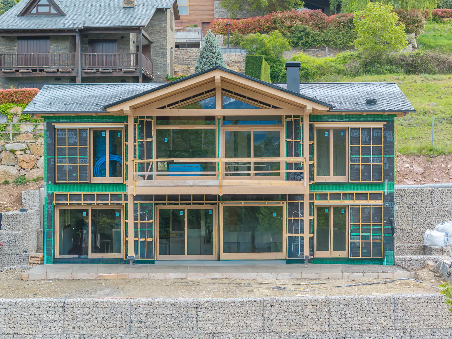 Two-story house under construction with wood framing and large windows. Black sheathing and green insulation are visible. Stone retaining wall in front.