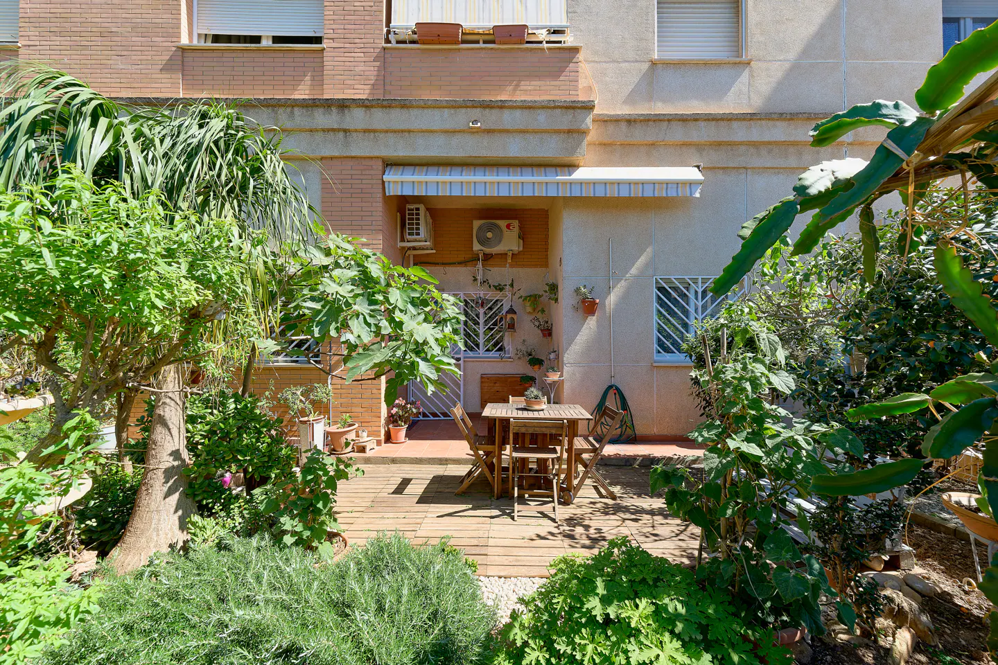 Outdoor patio with wooden deck, table, and chairs surrounded by lush greenery and a building facade in the background.