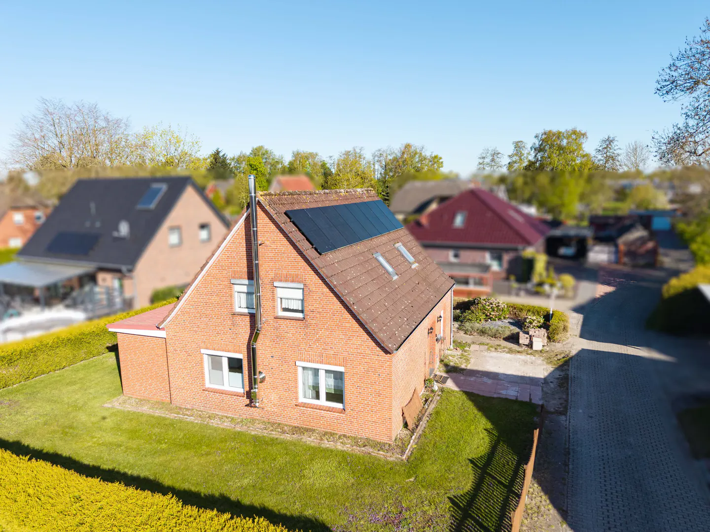 Aerial view of a red brick house with solar panels on the roof, surrounded by green lawn and other houses.