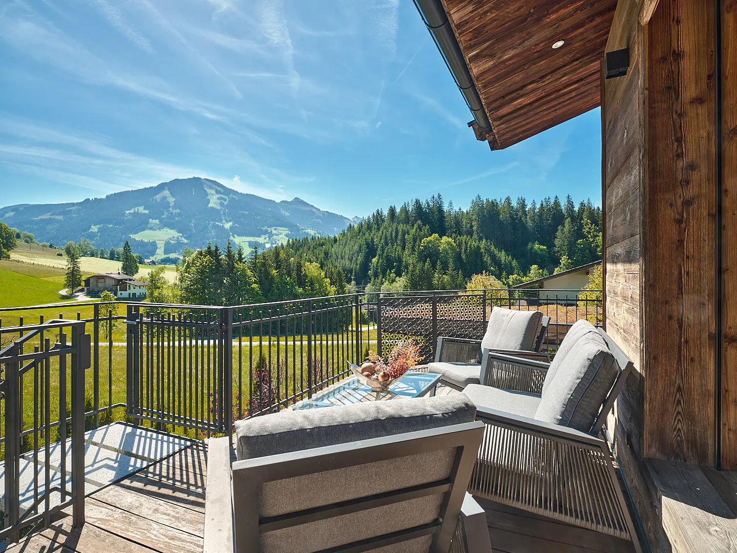 Balcony with gray cushioned chairs and glass table overlooking green hills and mountains under a blue sky.