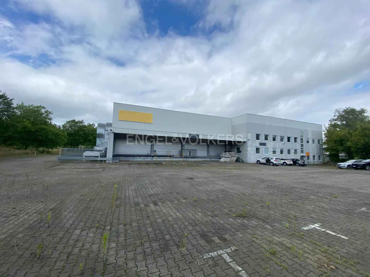 Gray warehouse building with loading docks and a large parking lot under a cloudy sky. Engel & Volkers logo on the building.