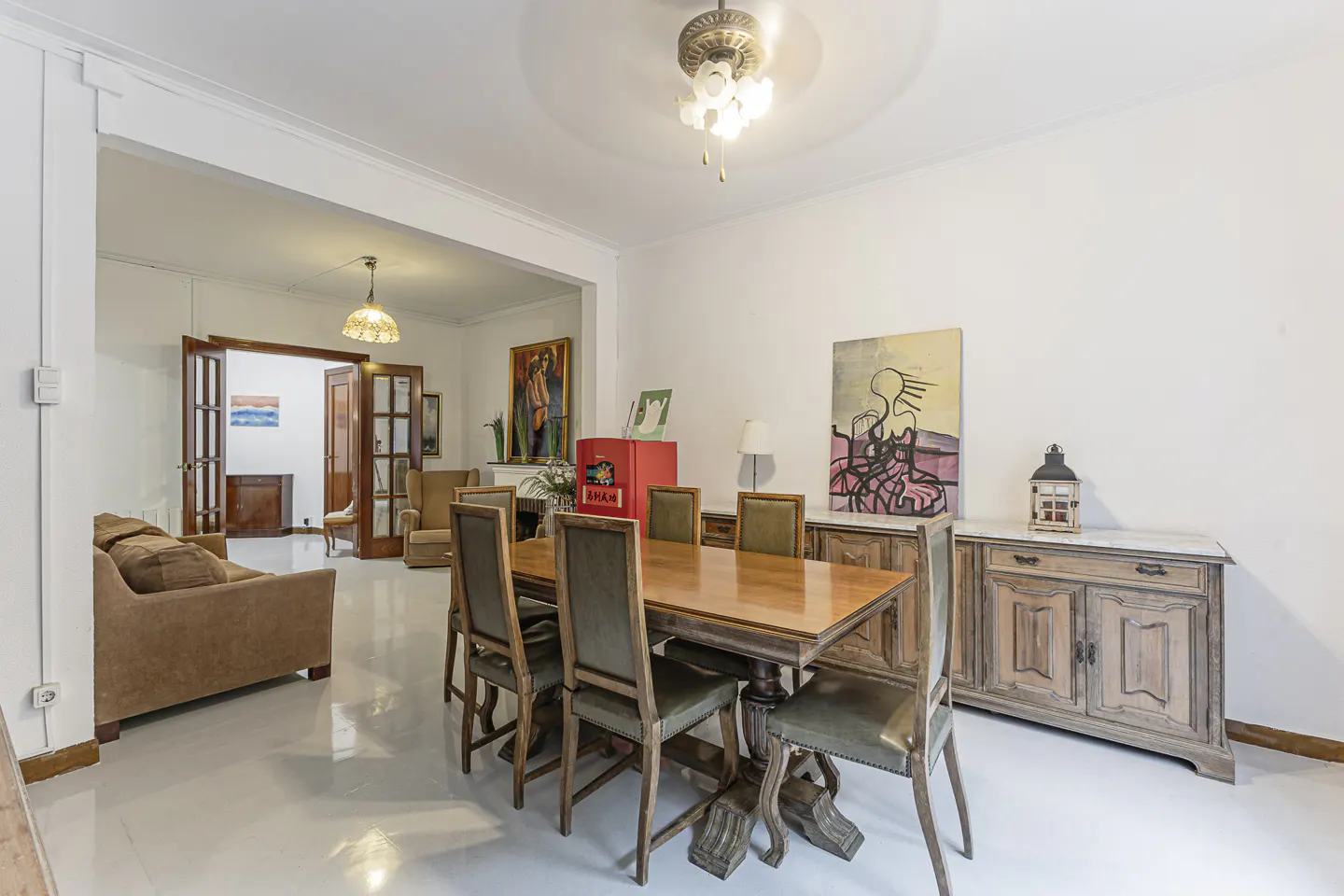 Bright dining room with a wooden table and six chairs, two wooden sideboards, and abstract art on the wall. A living room is visible through an archway.