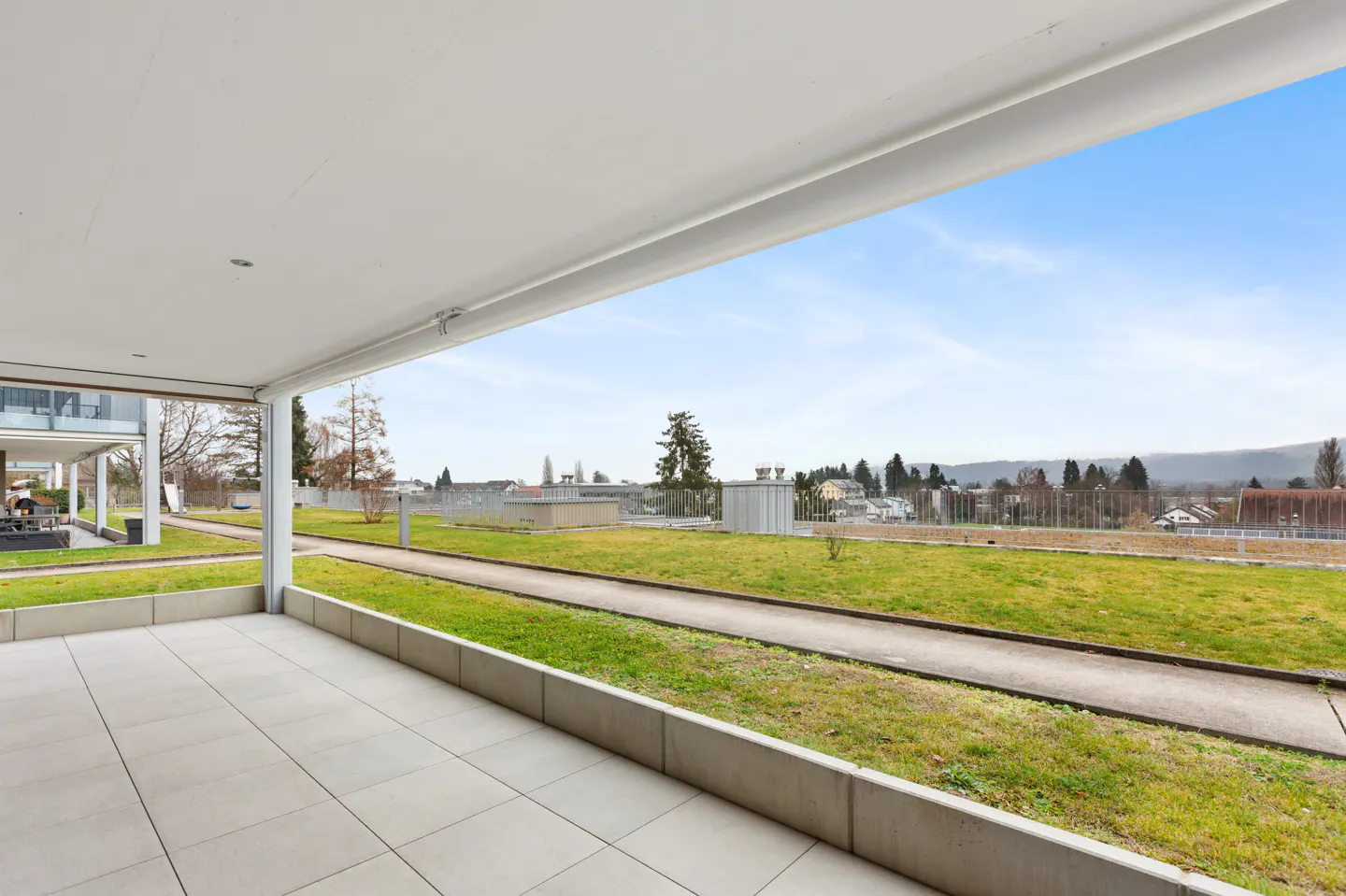 A covered patio with light gray tiles overlooks a green lawn and a distant town under a blue sky.