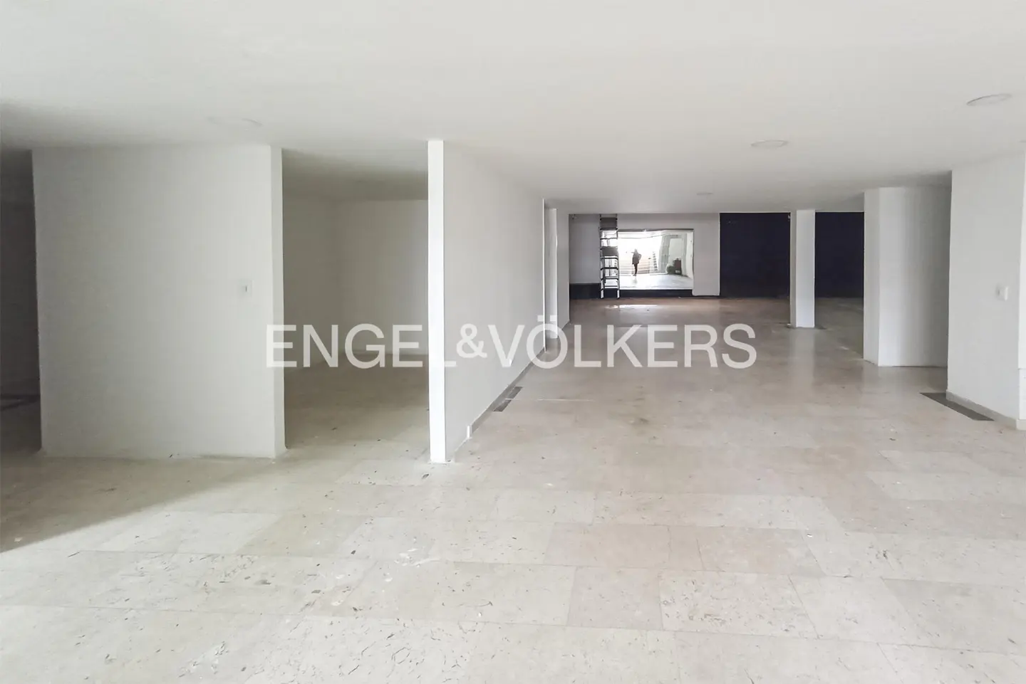 Unfurnished, open-concept space with white walls and light marble floors. Engel & Volkers logo is visible. Person in background.
