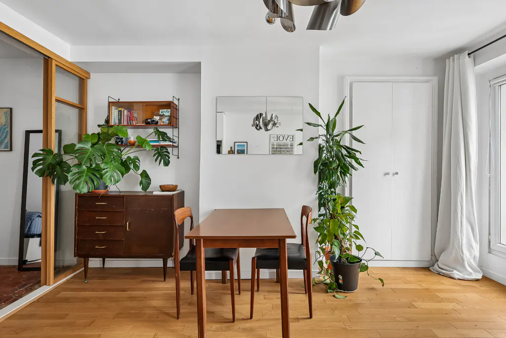 Dining area with wooden table, two chairs, vintage sideboard, wall shelving, large potted plants, and a wall mirror.