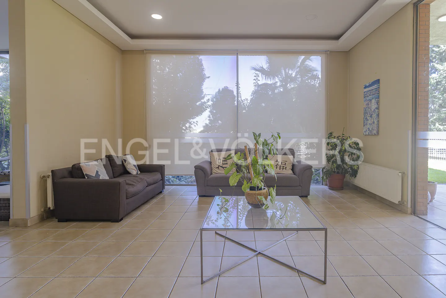 Living room with tile floors, two sofas, and a glass coffee table. Large windows with blinds overlook trees. Neutral color scheme.