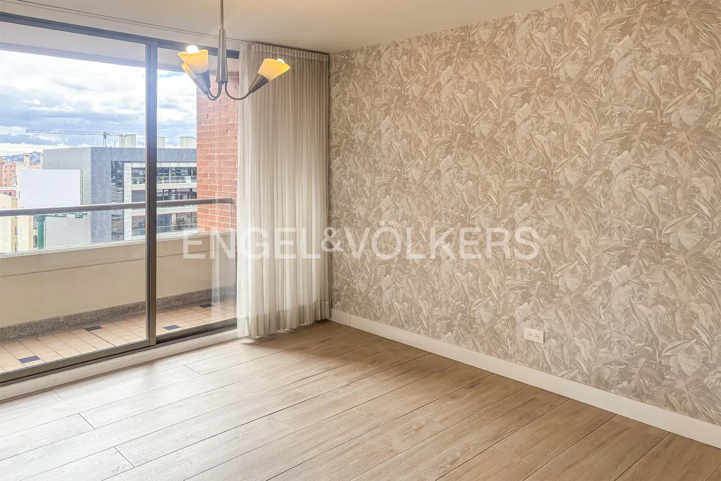 An empty room with light wood floors, a balcony with a city view, and leaf-patterned wallpaper. A chandelier hangs from the ceiling.