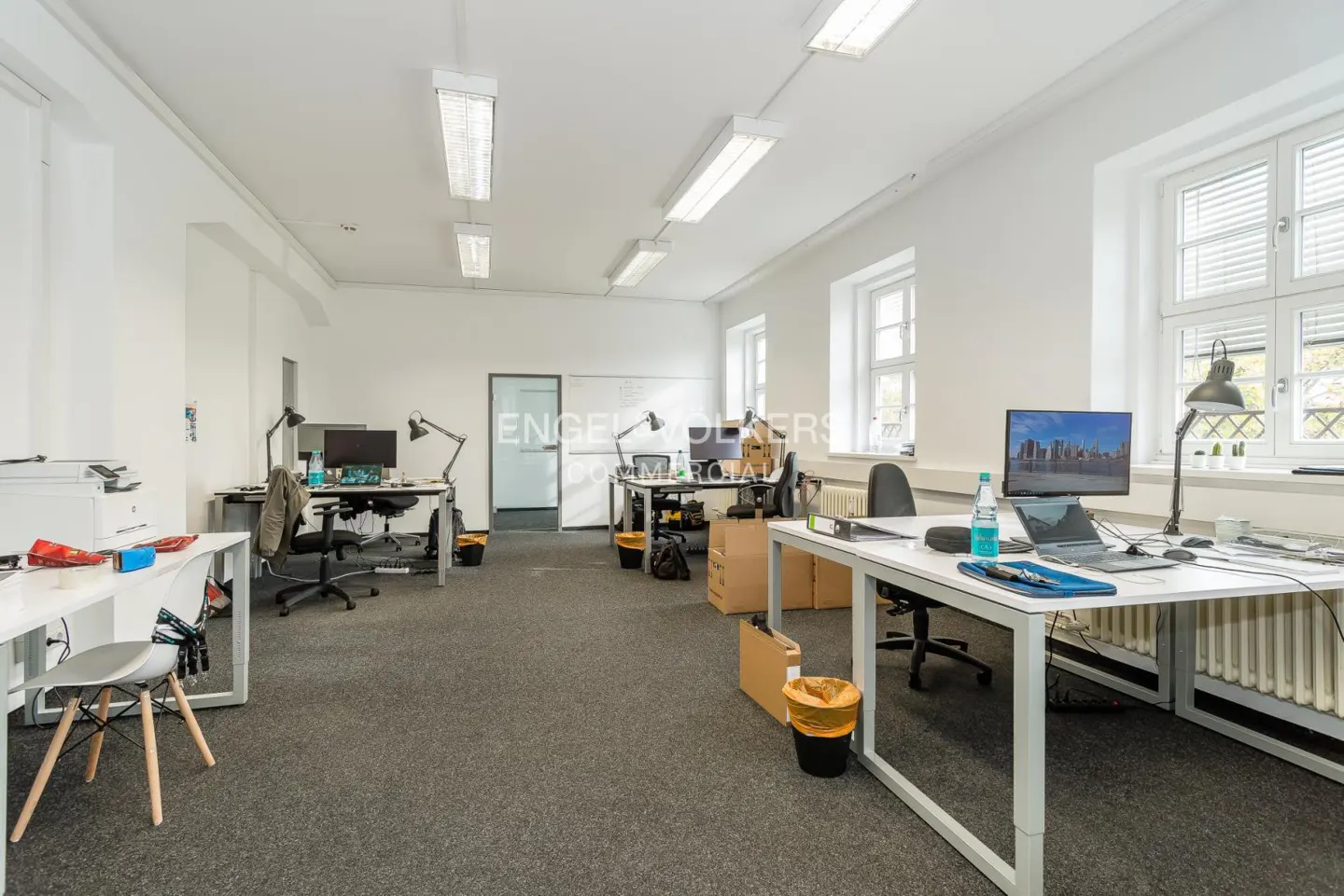 Bright office space with white walls, gray carpet, and desks with computers. Windows with blinds let in natural light.