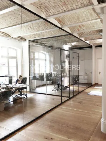 Office space with glass-walled rooms, wood floors, and brick arched ceiling. A person works at a desk in the background.