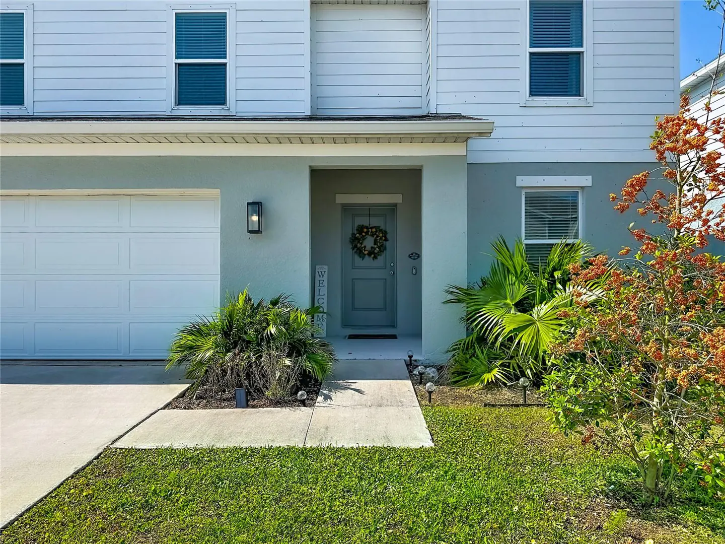 Two-story house with a white garage door, a gray front door with a wreath, and green landscaping.