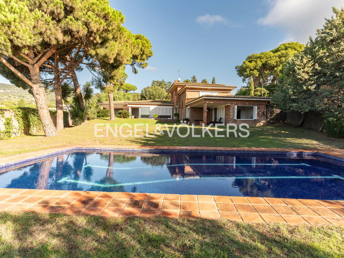 A wide shot of a brick house with a blue tiled pool in the foreground on a sunny day.