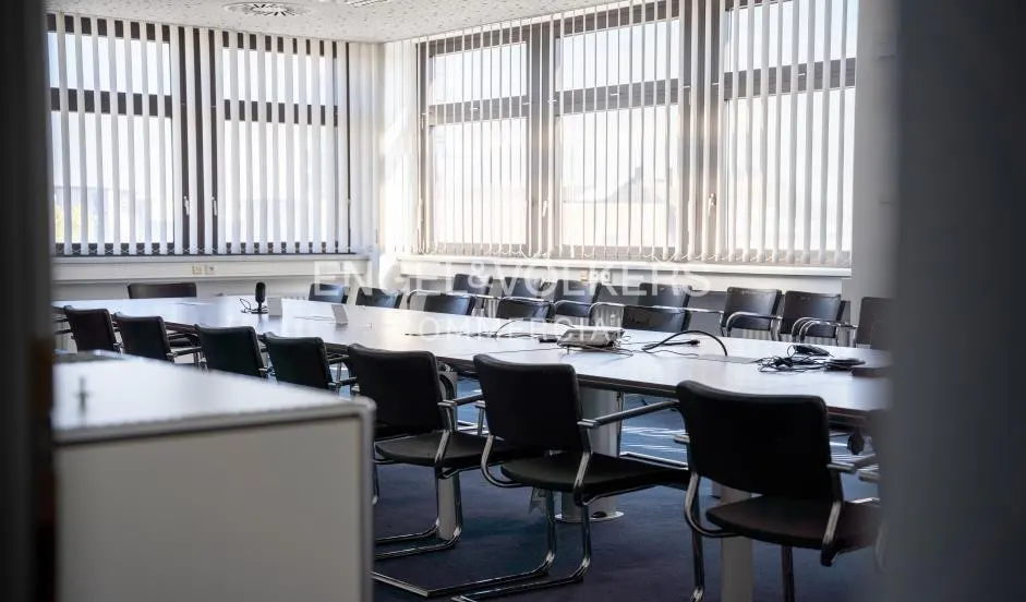 A bright conference room with a long table, black chairs, and large windows with vertical blinds.