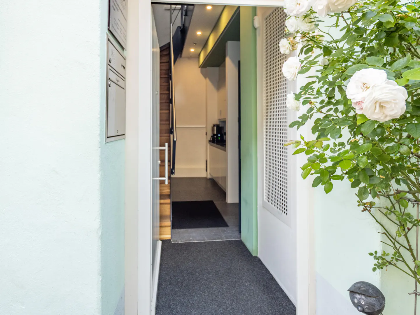 Open doorway with white trim and a light green wall. White roses are on the right. Inside, stairs lead up, and a kitchen area is visible.