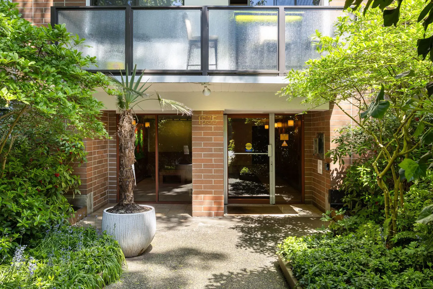 Building entrance with brick facade, glass doors, and transom windows. Lush greenery surrounds the entrance. A potted palm tree stands to the left.