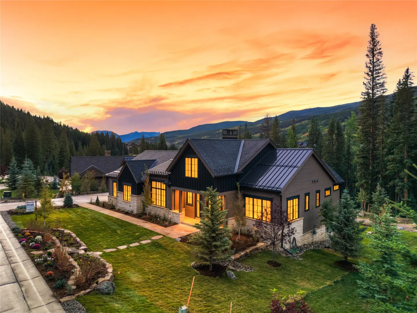 A modern two-story house with a green lawn and trees, set against a sunset sky and mountain backdrop.