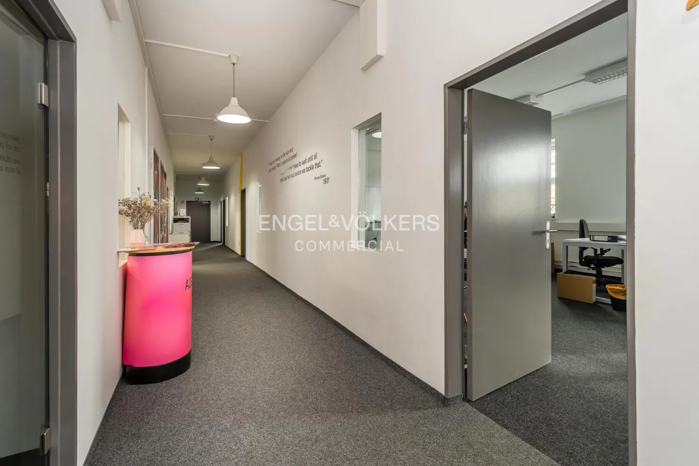 Office hallway with gray carpet, white walls, and pendant lights. A pink reception desk is on the left, and an open door reveals an office space.