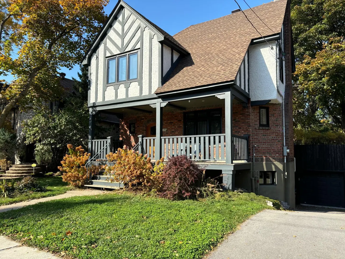 Two-story Tudor-style house with brick and stucco exterior, brown roof, and gray trim. A covered porch with railings is visible. Green lawn and trees surround the house.