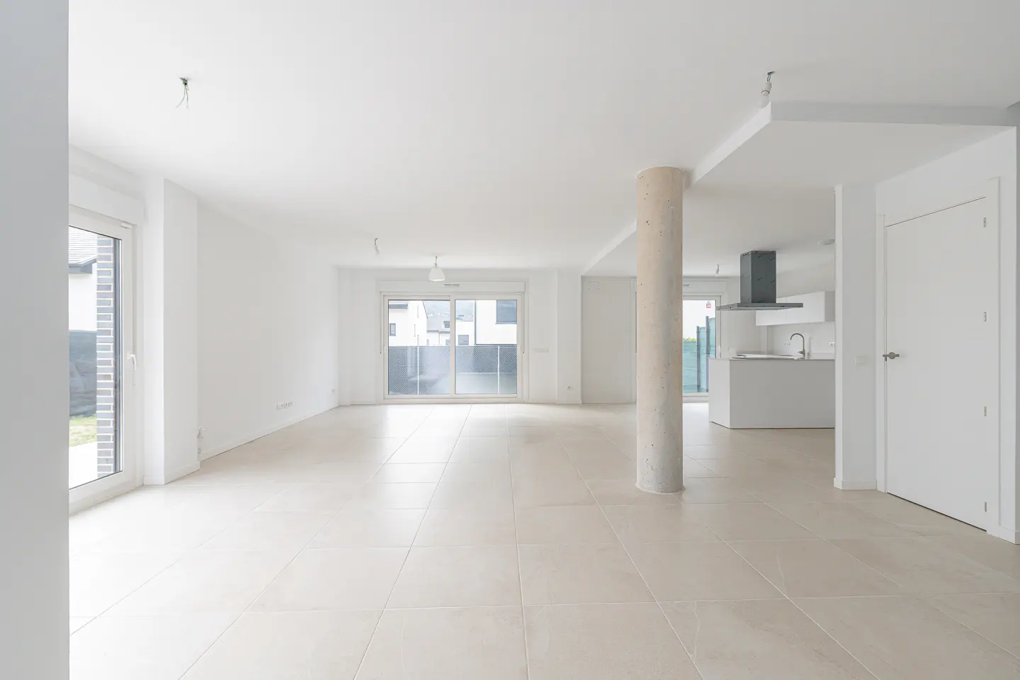 Bright, empty modern apartment with white walls, tile floors, and large windows. A concrete pillar separates the living area from the kitchen.