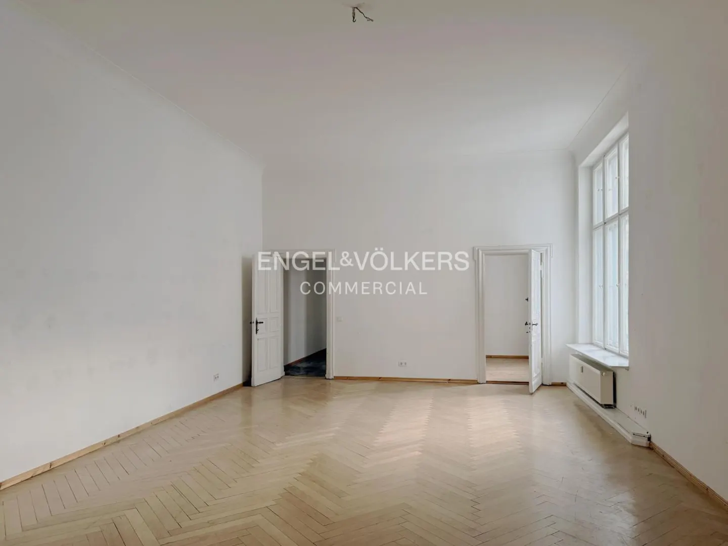 Bright, empty room with herringbone wood floors, white walls, and tall windows. Two open doorways lead to other rooms. Engel & Völkers logo.