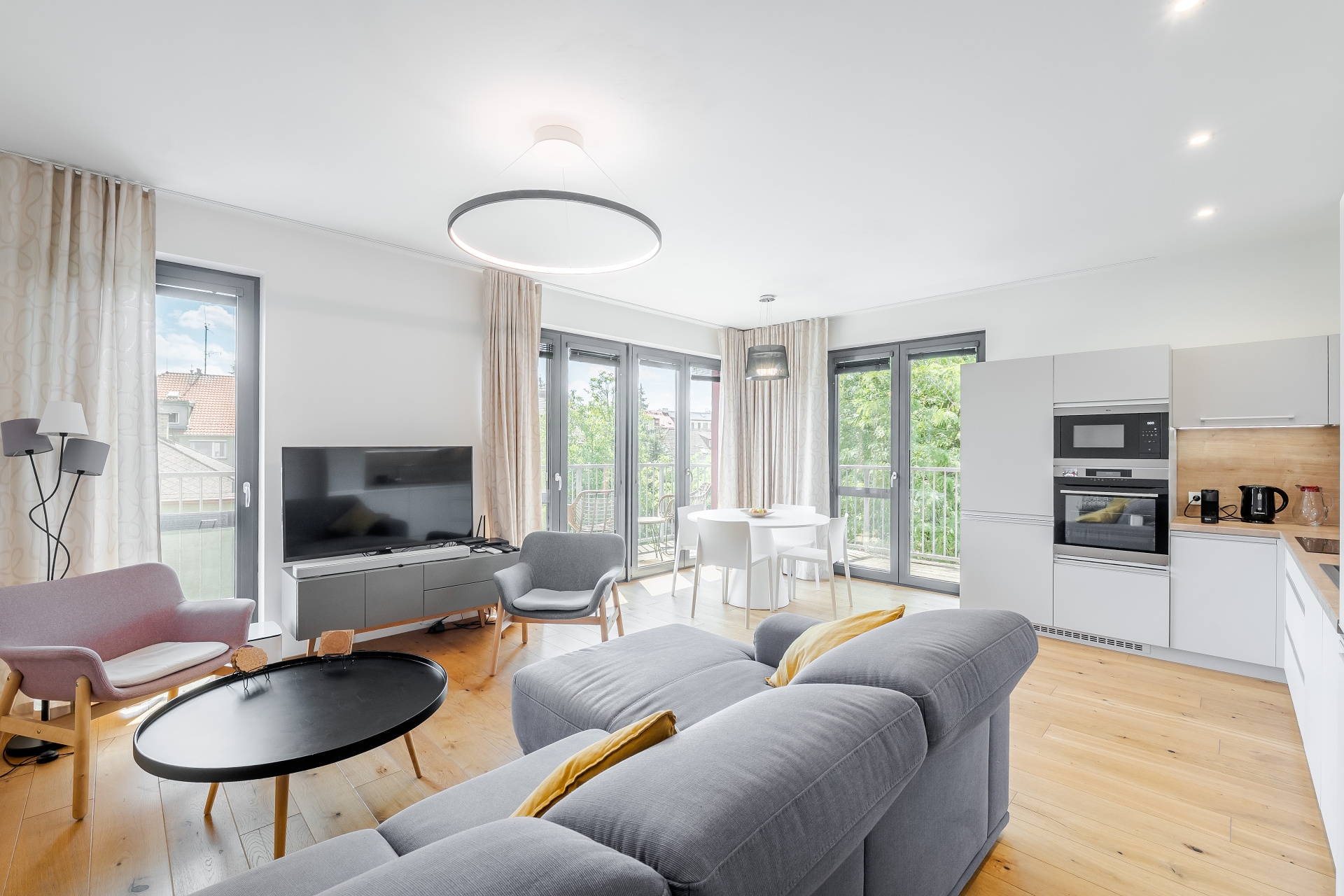 Modern open-plan living room with a grey sectional sofa, black coffee table, TV, dining area, and white kitchen cabinets.