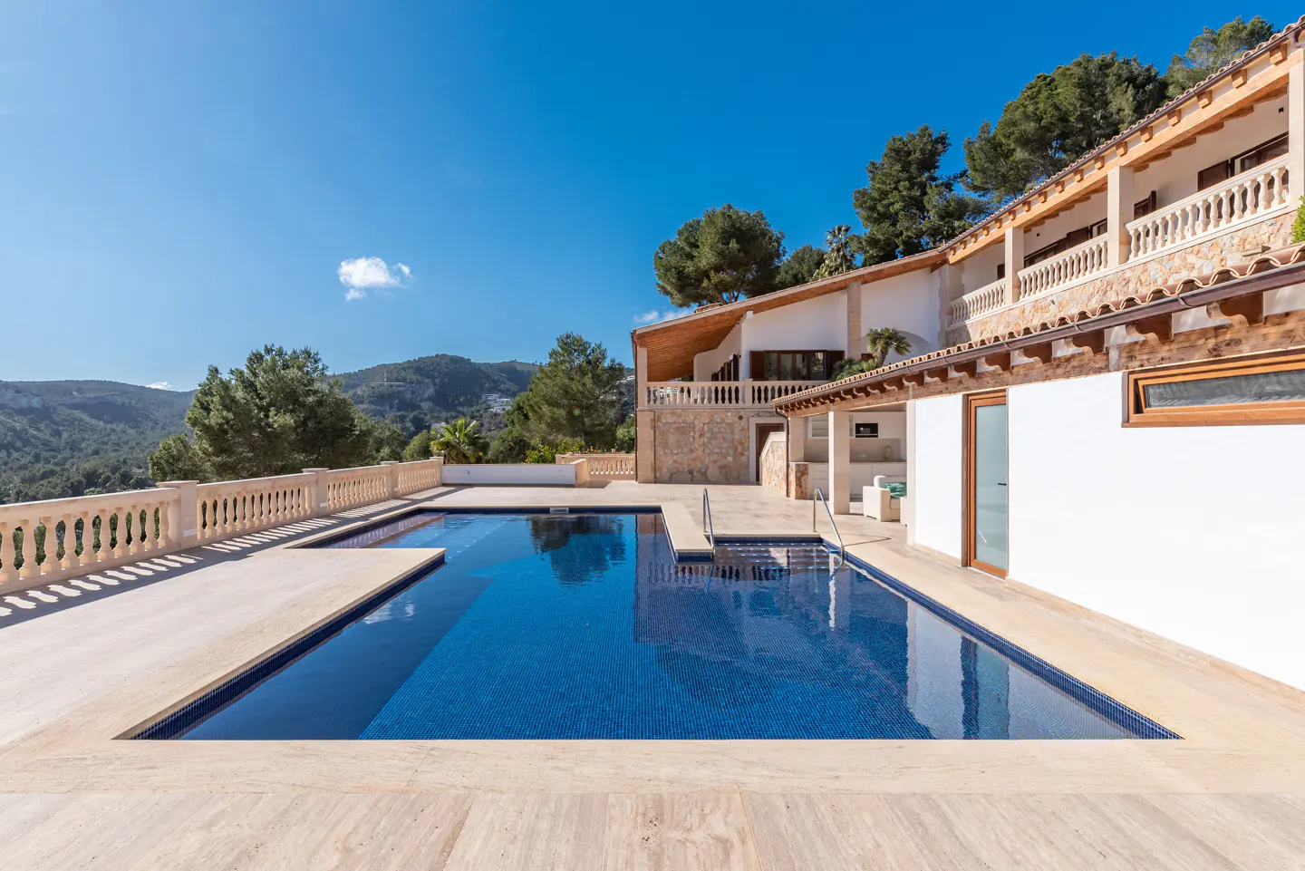 A blue tiled pool sits in front of a white house with balconies, under a clear blue sky. Trees and hills are in the background.