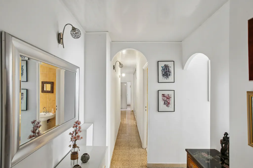 Hallway with white walls, arched doorways, and art. A large mirror reflects a bathroom. A long hallway extends into the distance.