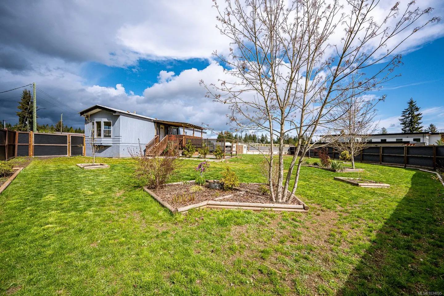 A wide shot of a light blue single-story home with a wooden porch and a large green lawn with garden beds.