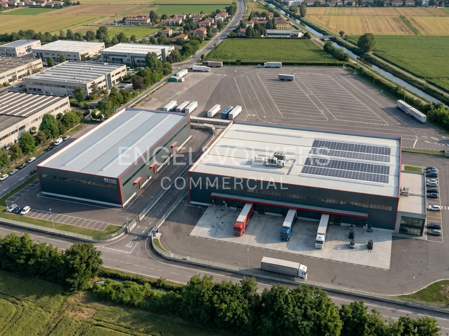 Aerial view of two gray commercial buildings with red accents, solar panels, and trucks at loading docks. Large parking lot and green fields surround the buildings.